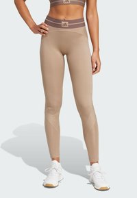 Beige højtaljede leggings med teksturerede detaljer og et brunt stribet bælte. Materialet virker strækbart og glat, egnet til sportstøj.