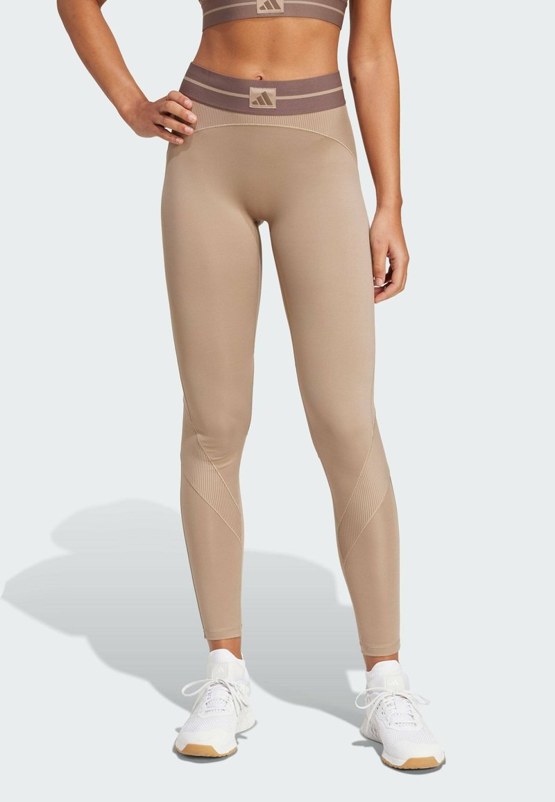 Beige højtaljede leggings med teksturerede detaljer og et brunt stribet bælte. Materialet virker strækbart og glat, egnet til sportstøj.