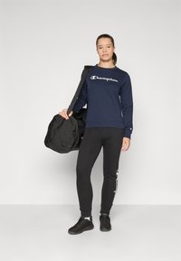 Sweatshirt marine avec le texte "Champion" blanc, associé à un pantalon noir ajusté. Un sac de sport noir et des baskets noires complètent le look.