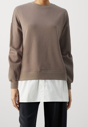 Personne portant un pull marron à manches longues superposé à une chemise blanche, associé à un pantalon noir, sur un fond clair uni.