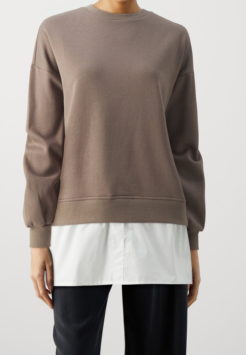 Personne portant un pull marron à manches longues superposé à une chemise blanche, associé à un pantalon noir, sur un fond clair uni.