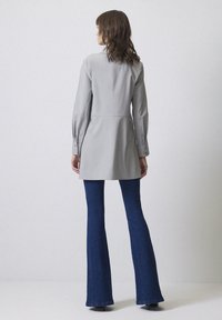 Grå tunika med långa ärmar, skräddarsydd passform och knappade manschetter, i kombination med utsvängda blå denimjeans. Enkel och ren design.