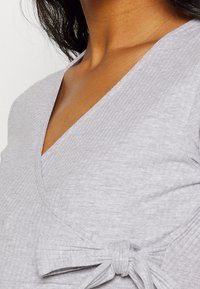 Närbild av en person klädd i en ljusgrå ribbad omlottopp med V-ringning och en tygrosett knuten vid midjan.