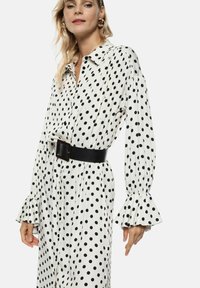 Abito camicia bianco con pois neri. Presenta un colletto, chiusura con bottoni, polsini elasticizzati e una cintura nera larga in vita.