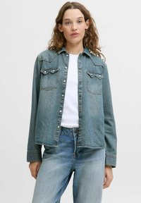 Giacca in denim azzurro chiaro con due tasche sul petto, chiusure a bottoni automatici e colletto classico. Il tessuto ha un aspetto strutturato.