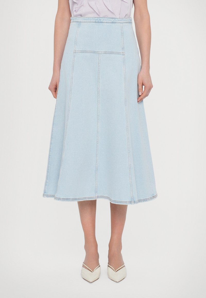 Kvinna som bär en ljusblå högmidjad denim midi kjol och vita spetsiga låga klackar, stående mot en neutral bakgrund.