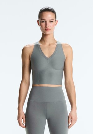 Kobieta z zaczesanymi do tyłu włosami, ubrana w szary sportowy biustonosz i wysoki stan legginsów, stoi na tle jednolitego jasnego tła.