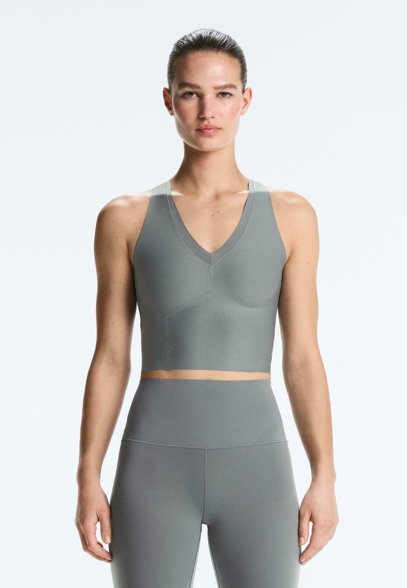 Frau mit zurückgebundenem Haar, die einen grauen Sport-BH und hoch taillierte Leggings trägt, steht vor einem schlichten hellen Hintergrund.