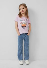 Rosa Baumwoll-T-Shirt mit einer Cartoon-Katze in einer Schüssel, umgeben von Blumen und einem Regenschirm. Kombiniert mit hellblauen Jeans und weißen Sneakers.