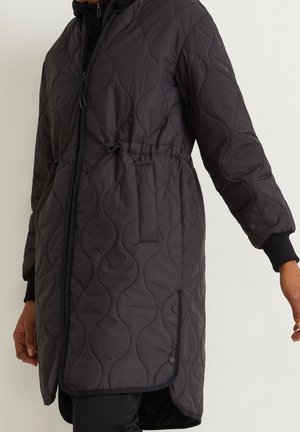Veste longue matelassée noire avec fermeture éclair, taille ajustable, poches latérales et boutons-pression à l'ourlet, portée sur un pantalon noir.