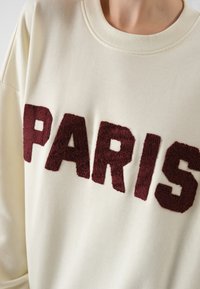 Krém színű pulóver, puha bordó színű "PARIS" felirattal. Kerek nyakkivágás és laza fazon, puha, textúrált anyaggal.