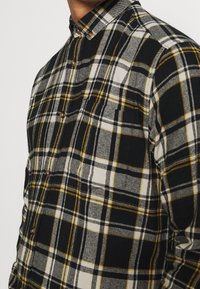 Camisa de cuadros en negro, blanco y amarillo, con cuello extendido, tejido texturizado y botones marrones, que presenta un bolsillo en el pecho.