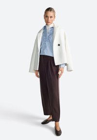 Cappotto bianco con texture bouclè e bottone nero, indossato sopra una camicia a righe azzurra chiara con volant, abbinato a pantaloni marrone scuro e ballerine.