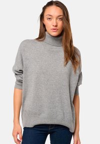 Jeune femme aux longs cheveux bruns portant un pull à col roulé gris ample et un jean bleu foncé devant un fond blanc uni.