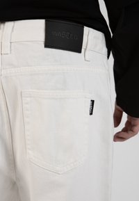 Los pantalones de mezclilla blancos cuentan con un bolsillo trasero con una etiqueta negra inscrita con la palabra "WASTED". La tela tiene una textura suave y un ajuste estructurado.