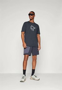 Vyras, vilkintis juodą grafikos piešiniu marškinėlį, tamsias šortus su pleistru, juodas kojines, baltais sportiniais bateliais ir dideliais juodais saulės akiniais, stovintis prieš šviesią fono.