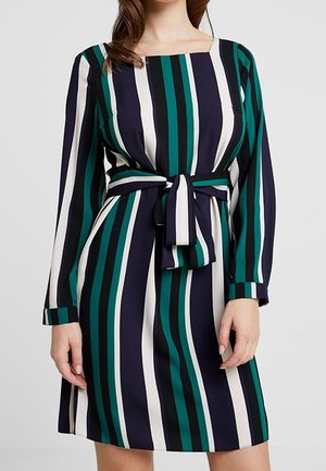 Femme portant une robe longue jusqu'aux genoux à manches longues avec des rayures verticales vertes, bleu marine, noires et blanches et une ceinture nouée à la taille.