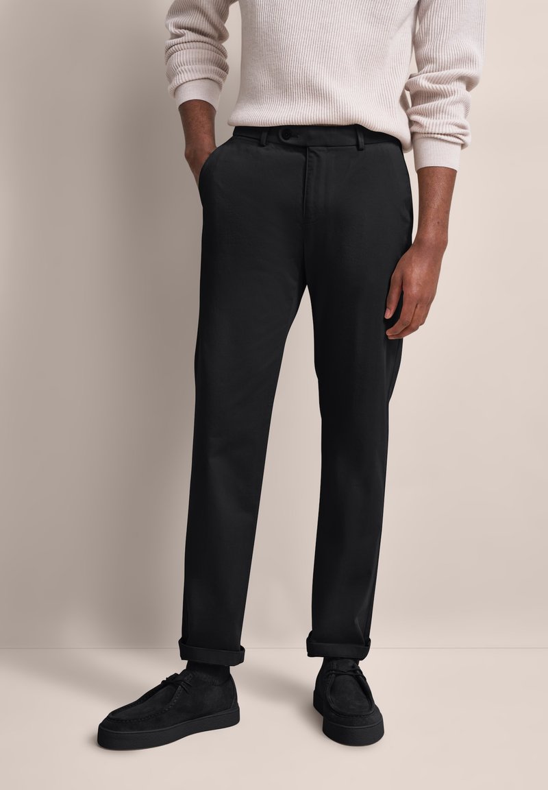 Mężczyzna w czarnych spodniacjach z mankietami, czarnych butach i beżowej, prążkowanej bluzie z długim rękawem, stojący na neutralnym tle.