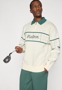 Pull-over blanc en tricot avec des accents verts, doté d'un col zippé et d'un motif à carreaux. Un club de golf est tenu dans la main du mannequin.