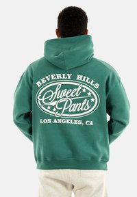 Sudadera verde con un gran gráfico blanco en la parte de atrás que presenta "Beverly Hills Sweet Pants Los Ángeles, CA" y estrellas decorativas.