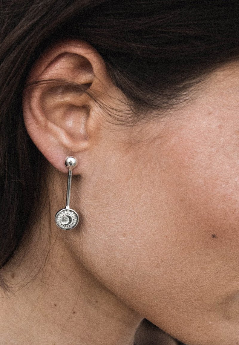 Söljdängande örhängen med en rund skiva med solstrålemönster och en slät sfärisk topp, hängande från en tunn stav.