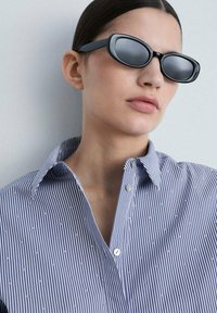 Black oval sunglasses with tinted lenses, worn with a blue and white striped button-up shirt featuring small white dots.