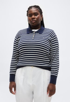 Frau mit geflochtenem Haar, die ein marineblau-weiß gestreiftes langärmliges Poloshirt und weiße Hose trägt, steht vor einem einfarbigen Hintergrund.