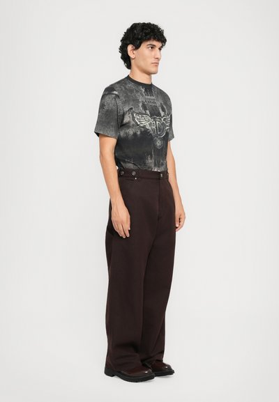 Man with curly hair stands facing right, wearing a black graphic t-shirt, dark brown baggy pants, and dark brown shoes on a white background.