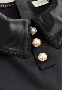 Blusa nera con colletto appuntito in raso rifinito con pizzo smerlato e tre grandi bottoni di perle con bordatura dorata sulla patta.