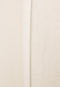Kabelstickad tröja i off-white med ribbad textur på vänster sida och slätt tyg på högra sidan, med en central sömdetalj.