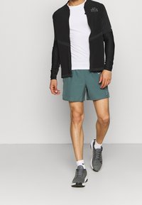 Man met een zwarte ritsjack, een wit T-shirt, teal hardloopshorts, witte sokken en grijze hardloopschoenen die voorwaarts loopt.
