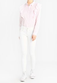 Blusa a righe rosa chiaro con dettagli arricciati sulla spalla sinistra, chiusura con bottoni sul davanti, abbinata a jeans skinny bianchi e sneakers bianche.