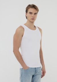 Top in cotone bianco a canotta con scollatura rotonda e texture a costine, abbinato a jeans azzurri chiari. Il modello sta in piedi con le mani in tasca.