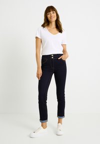 Jeans azul oscuro de talle alto con dobladillo desgastado, que presentan dos bolsillos frontales, detalles de botones dorados y puños enrollados, combinados con una camiseta blanca.