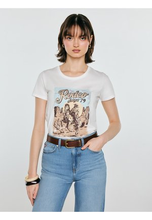 Młoda kobieta w białej koszulce z grafiką "Rodeo days '79", niebieskich dżinsach, brązowym pasku i kolczykach-kołach, pozująca z ręką w kieszeni.