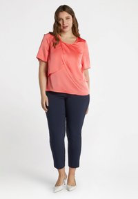 Korallfarbendes Satinbluse mit kurzen Ärmeln, drapiertem Design und besticktem Ausschnitt; kombiniert mit marineblauen, figurbetonten Hosen und weißen Absatzschuhen.