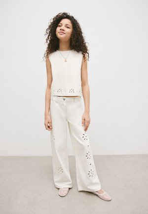 Meisje met krullend haar draagt een mouwloos wit topje en witte broek met bloemenvormige uitsnijdingen, staand op een grijze vloer tegen een effen muur.