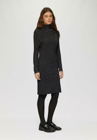 Schwarzes langärmliges Kleid mit hohem Kragen, das eine funkelnde Textur aufweist. Tragen mit schwarzen Strumpfhosen und besetzten schwarzen Schuhen.