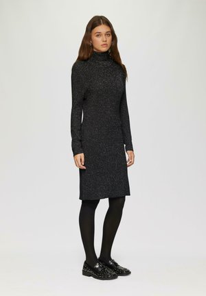 Schwarzes langärmliges Kleid mit hohem Kragen, das eine funkelnde Textur aufweist. Tragen mit schwarzen Strumpfhosen und besetzten schwarzen Schuhen.
