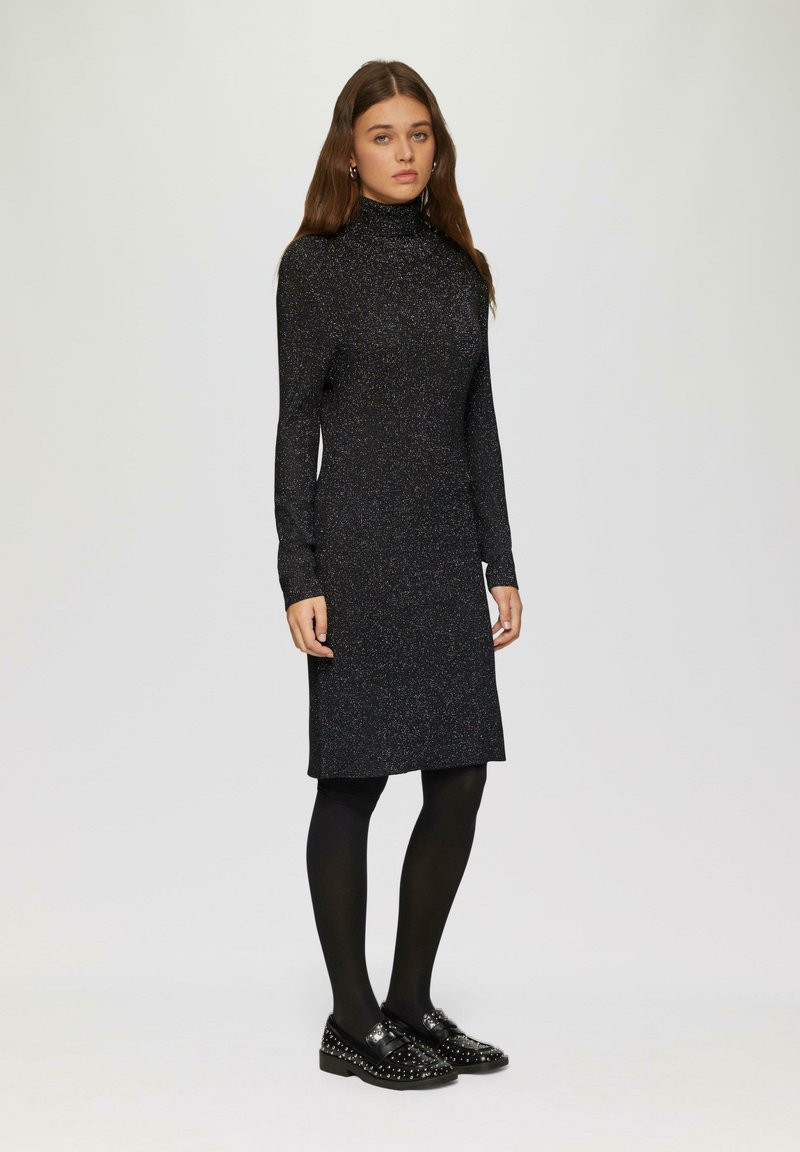 Schwarzes langärmliges Kleid mit hohem Kragen, das eine funkelnde Textur aufweist. Tragen mit schwarzen Strumpfhosen und besetzten schwarzen Schuhen.
