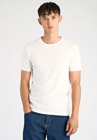 T-shirt bianca in cotone con scollo rotondo e maniche corte, abbinata a jeans denim blu scuro, con un design semplice e logo discreto.