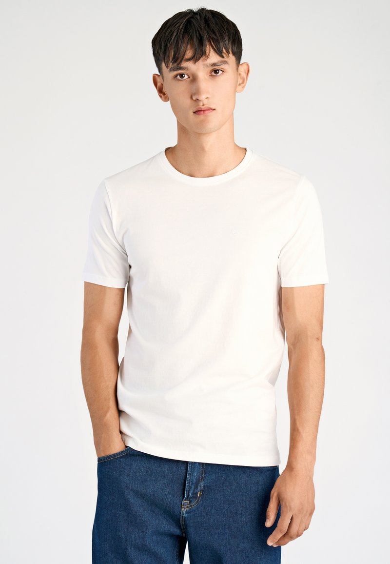 T-shirt bianca in cotone con scollo rotondo e maniche corte, abbinata a jeans denim blu scuro, con un design semplice e logo discreto.