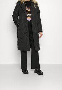 Zwarte gewatteerde lange jas met een capuchon, voorzien van drukknoopsluitingen en diagonale stiksels. Gepaard met zwarte wijde broek en stevige zwarte schoenen.