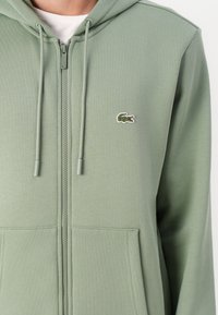 Sudadera verde con cremallera de suave tejido, con un bolsillo tipo canguro, que presenta un pequeño logo de cocodrilo bordado en el lado izquierdo del pecho.
