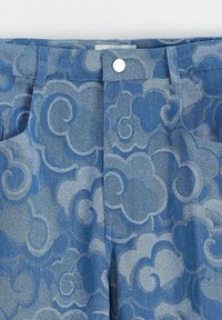 Pantalons en denim avec un motif nuage bleu et bleu clair, dotés d'une fermeture à bouton sur le devant et de poches latérales, mettant en avant un tissu texturé.