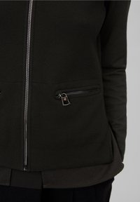 Black zip-up jacket made from textured fabric, featuring two side zip pockets and ribbed cuffs. Design is fitted with a straight hem.