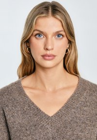 Pulover tricotat maro cu decolteu în V, având un model texturat. Modelul are păr blond deschis și poartă cercei rotunzi.