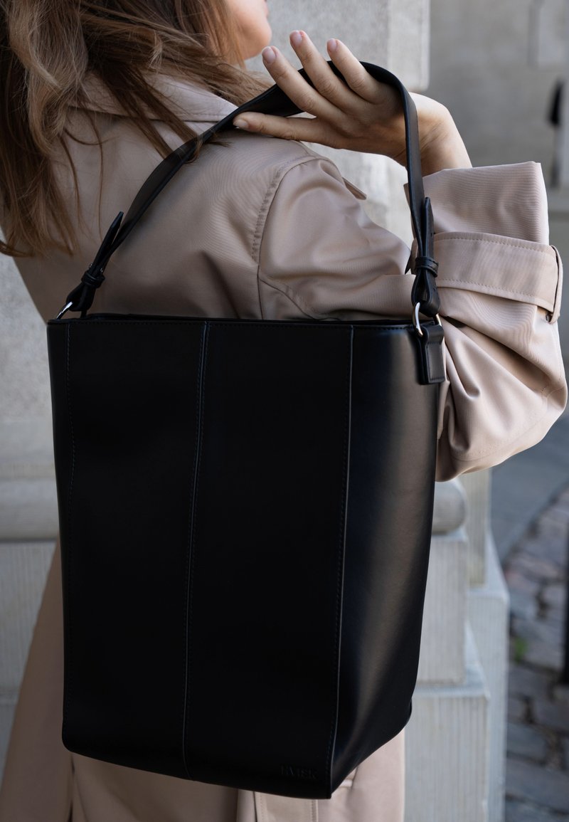 Femme portant un trench-coat beige avec un grand sac à main en cuir noir sur l'épaule, en extérieur près d'un mur en pierre.