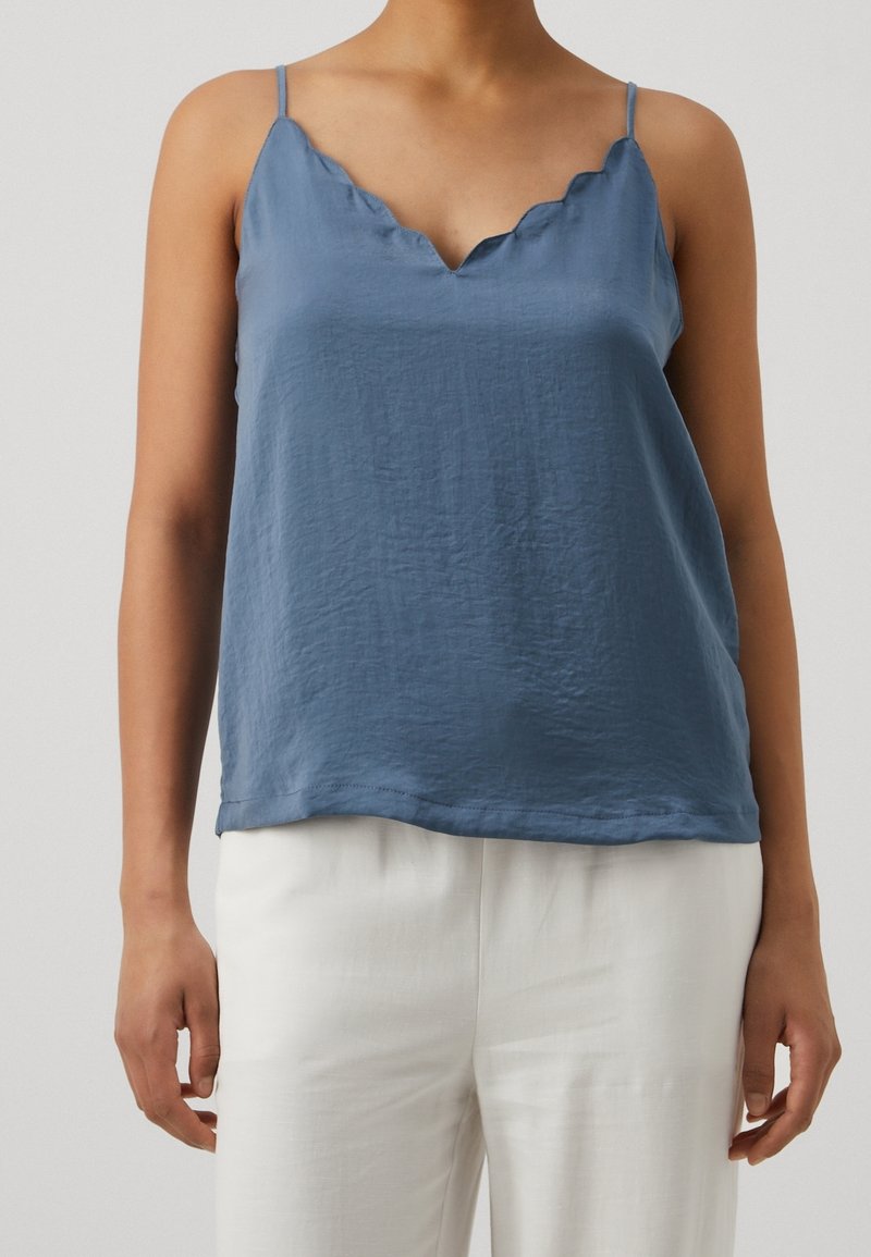 Personne portant un haut bleu sans manches à bords festonnés et un pantalon blanc, debout avec les bras détendus le long du corps devant un fond uni.