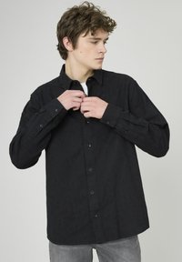 Chemise noire à boutons avec un motif texturé, manches longues et boutons noirs, associée à un pantalon gris.